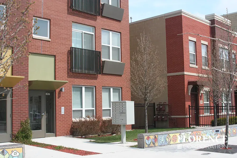 Single vital&trade; cluster box unit in postal gray installed outside modern brick multifamily housing apartment building with mosaic mural along short wall lining sidewalk
