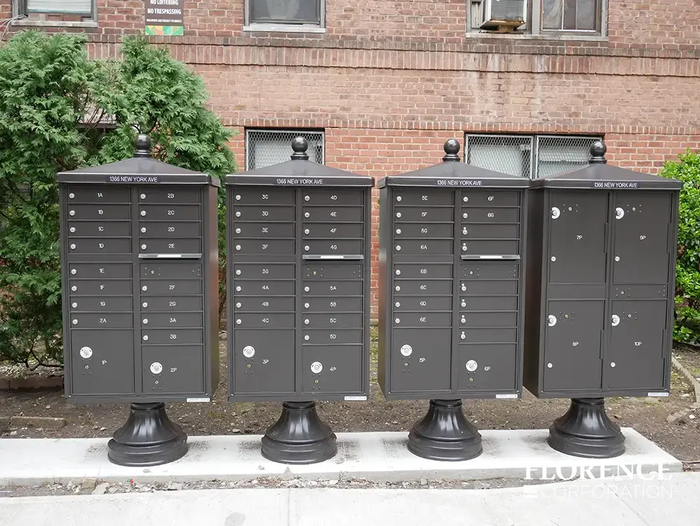 valiant&trade; outdoor parcel locker and vital&trade; cluster box units with vogue&trade; accessories all in dark bronze  installed on concrete slab in front of red brick apartment building near green trees and urban landscaping