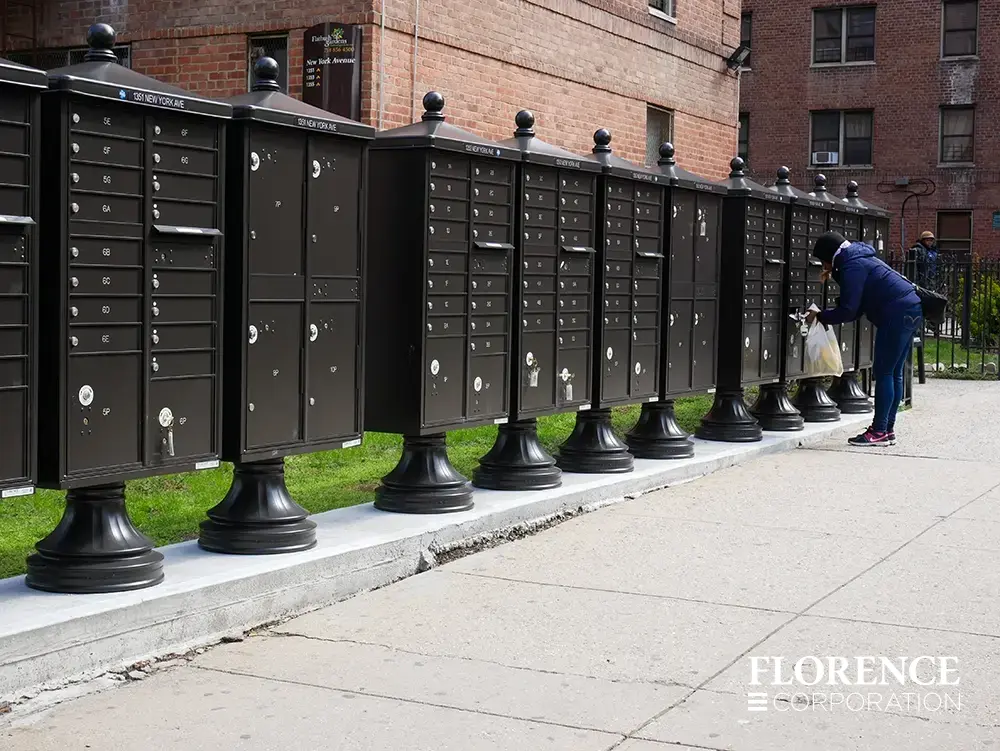 valiant&trade; outdoor parcel locker and vital&trade; cluster box units all with vogue&trade; accessories in dark bronze installed on concrete slab in brooklyn nyc apartment buildings with woman in sneakers retrieving her mail from one cbu