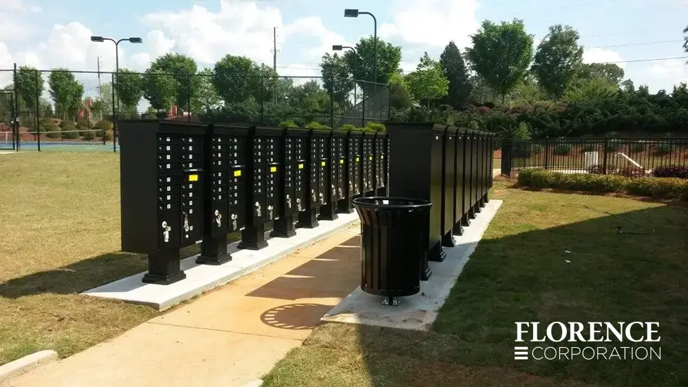 vogue&trade; accessories on vital&trade; cluster box units in black installed on white concrete slabs on both sides of the a sidewalk near recreation center park