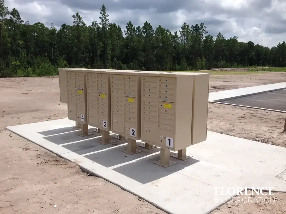 multiple vital&trade; cluster box units in sandstone installed back-to-back on wide concrete slab surrounded by dirt ground near parking lot still in development with green forest in the background and cloudy sky