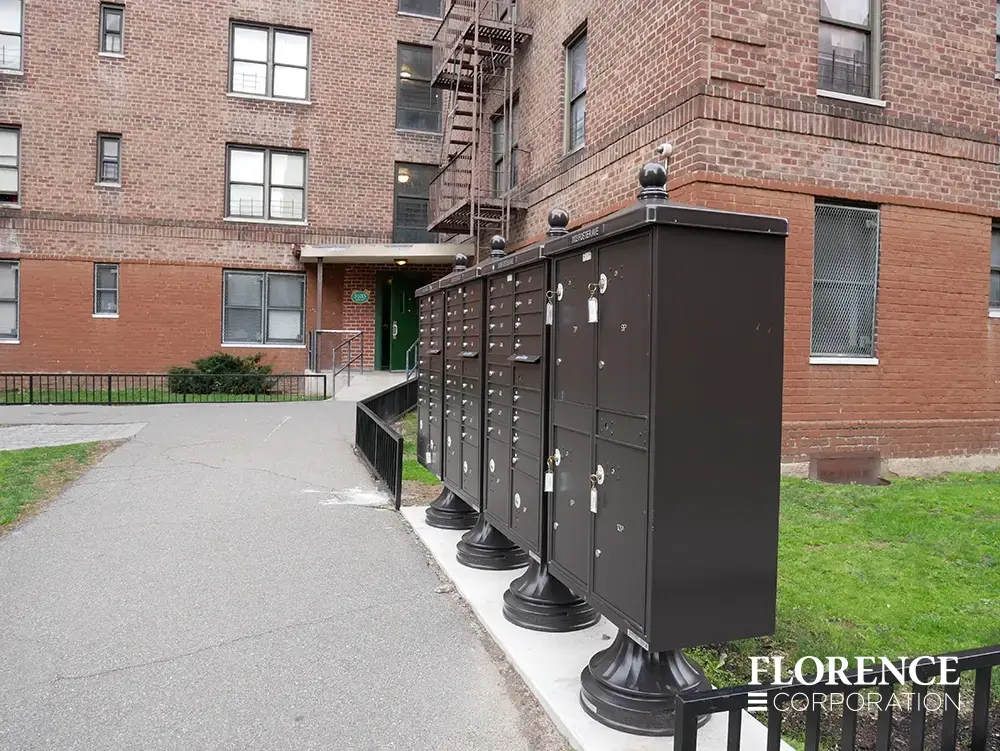 valiant-1590-OPL-series_009 valiant™ outdoor parcel locker and vital™ cluster box units with vogue™ decorative accessories all in dark bronze installed on concrete strip along sidewalk and grassy lawn in front of red brick apartment building
