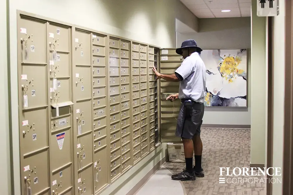 versatile-4C-indoor-install_022 Recessed mount versatile™ 4C mailboxes and parcel lockers in sandstone installed in a hallway of a modern multifamily apartment building with floral painting in background and usps postal carrier delivering mail with master access door open