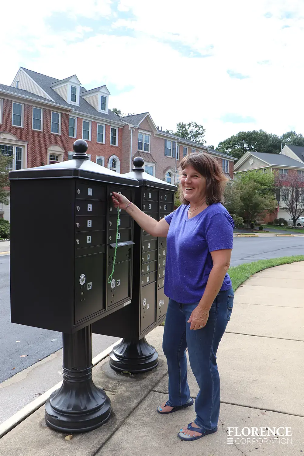 vogue™ accessories on vital™ cluster box units in dark bronze installed along sidewalk in town home community with smiling woman opening her mailbox to retrieve her mail